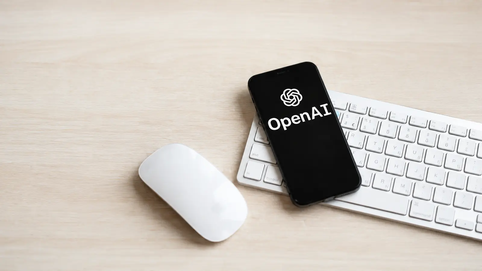 OpenAI logo displayed on a smartphone placed over a white keyboard on a light wooden desk
